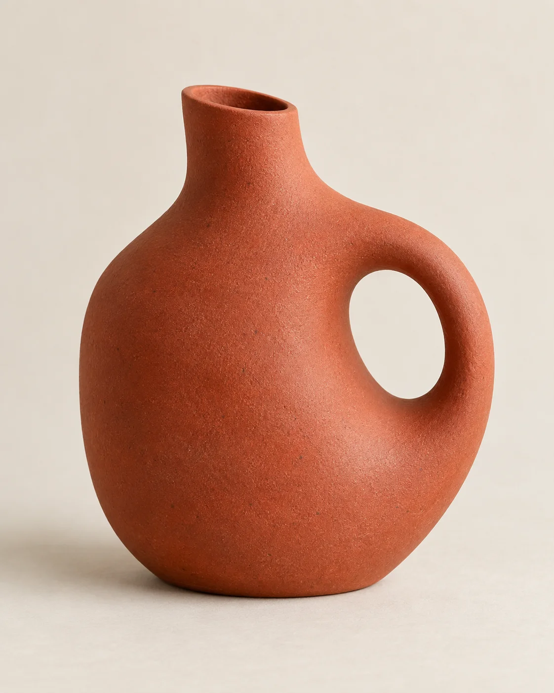 Terracotta ceramic vase with a circular handle on a warm ivory background.